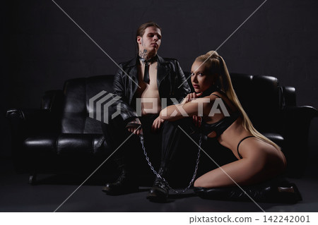 Dominant tattooed man and submissive woman in leather with chain on black couch Dominant tattooed man and submissive woman in leather with chain on black couch 142242001