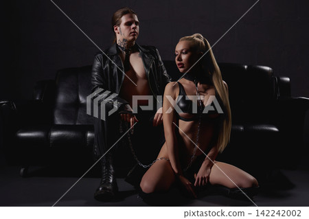 Edgy couple in black leather with chains posing in dark studio. Man with tattoos holds leash on kneeling blonde woman Edgy couple in black leather with chains posing in dark studio. Man with tattoos holds leash on kneeling blonde woman 142242002
