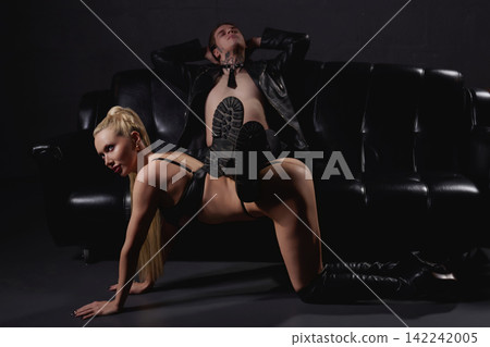 Edgy couple in black leather lingerie and jacket posing provocatively on dark sofa Edgy couple in black leather lingerie and jacket posing provocatively on dark sofa 142242005