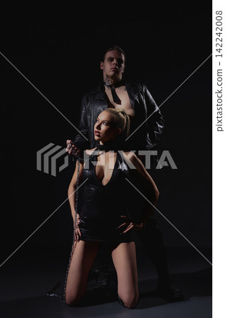 Dominant man in leather holds kneeling woman on chain against black background Dominant man in leather holds kneeling woman on chain against black background 142242008