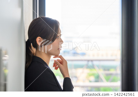 A female employee in a suit is deep in thought by the window. A female employee in a suit is deep in thought by the window. 142242561