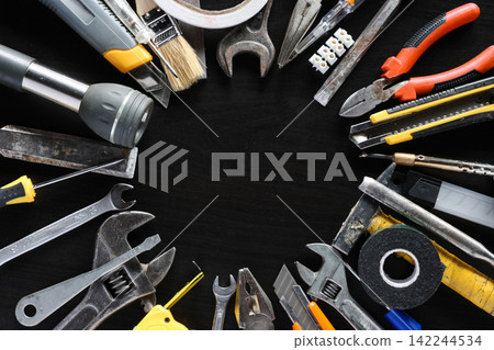 Assorted hand tools and repair supplies arranged around empty center on dark workshop surface Assorted hand tools and repair supplies arranged around empty center on dark workshop surface 142244534