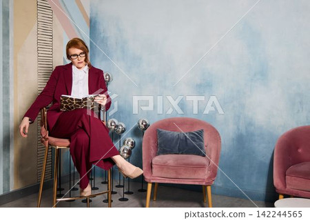Stylish woman in burgundy attire holding teacup while seated confidently Stylish woman in burgundy attire holding teacup while seated confidently 142244565