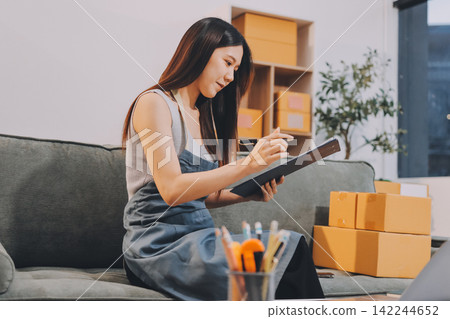 Startup SME small business entrepreneur of freelance Asian woman wearing apron using laptop and box to receive and review orders online to prepare to pack sell to customers, online sme business ideas. Startup SME small business entrepreneur of freelance Asian woman wearing apron using laptop and box to receive and review orders online to prepare to pack sell to customers, online sme business ideas. 142244652