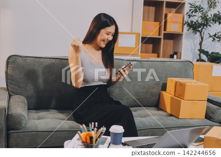 Startup SME small business entrepreneur of freelance Asian woman wearing apron using laptop and box to receive and review orders online to prepare to pack sell to customers, online sme business ideas. 142244656