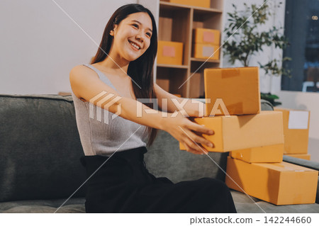 Startup SME small business entrepreneur of freelance Asian woman wearing apron using laptop and box to receive and review orders online to prepare to pack sell to customers, online sme business ideas. Startup SME small business entrepreneur of freelance Asian woman wearing apron using laptop and box to receive and review orders online to prepare to pack sell to customers, online sme business ideas. 142244660