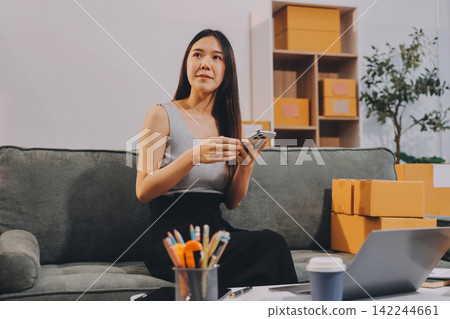 Startup SME small business entrepreneur of freelance Asian woman wearing apron using laptop and box to receive and review orders online to prepare to pack sell to customers, online sme business ideas. Startup SME small business entrepreneur of freelance Asian woman wearing apron using laptop and box to receive and review orders online to prepare to pack sell to customers, online sme business ideas. 142244661
