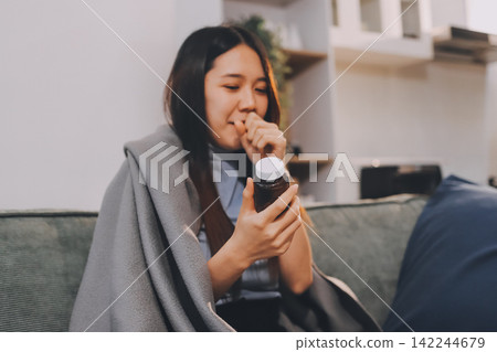 sick asian young woman sneezing into tissue paper while covered with a blanket and sitting on sofa in the living room sick asian young woman sneezing into tissue paper while covered with a blanket and sitting on sofa in the living room 142244679
