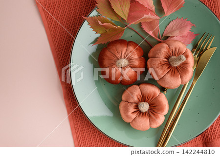 Three plush velvet pumpkins and autumnal leaves arranged on a teal plate with gold cutlery for a festive seasonal table setting concept. Three plush velvet pumpkins and autumnal leaves arranged on a teal plate with gold cutlery for a festive seasonal table setting concept. 142244810