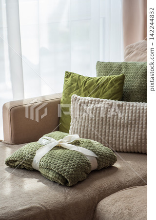 Cozy green blanket tied with white ribbon resting on comfortable sofa with plush pillows Cozy green blanket tied with white ribbon resting on comfortable sofa with plush pillows 142244832