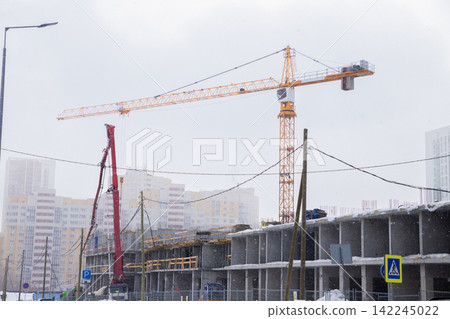 Yellow crane on construction site with concrete building frame under snowy sky. Urban development project with industrial machinery during winter. Infrastructure industry and architecture. Yellow crane on construction site with concrete building frame under snowy sky. Urban development project with industrial machinery during winter. Infrastructure industry and architecture. 142245022