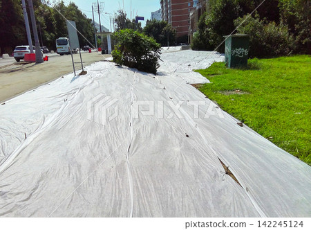 White landscape fabric covers the ground near an urban road to prevent weed growth in the city district. White landscape fabric covers the ground near an urban road to prevent weed growth in the city district. 142245124