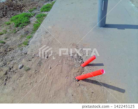Two orange plastic pipes stick out from a sandy ground near a grey sidewalk post on a sunny summer day. Two orange plastic pipes stick out from a sandy ground near a grey sidewalk post on a sunny summer day. 142245125