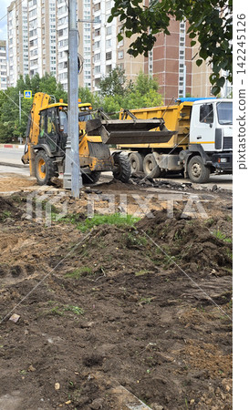 Yellow backhoe loader digs ground near white dump truck at urban construction site with residential houses. Yellow backhoe loader digs ground near white dump truck at urban construction site with residential houses. 142245126