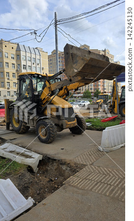 A yellow backhoe loader standing on a city street near residential buildings during a road repair project. A yellow backhoe loader standing on a city street near residential buildings during a road repair project. 142245130