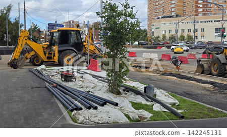 Yellow backhoe loader parked on urban road construction site during daytime with pipes and building machines. Yellow backhoe loader parked on urban road construction site during daytime with pipes and building machines. 142245131