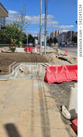 A construction site on a city street with red plastic barriers and concrete curbs during a sunny daytime. 142245134
