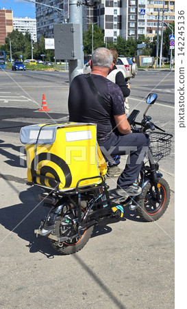 A middle aged caucasian man rides an electric scooter with a yellow delivery bag on a sunny city street. A middle aged caucasian man rides an electric scooter with a yellow delivery bag on a sunny city street. 142245136