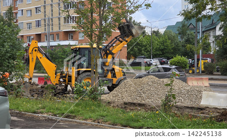 A yellow backhoe loader works on a city street near residential buildings, digging up ground for repair. A yellow backhoe loader works on a city street near residential buildings, digging up ground for repair. 142245138