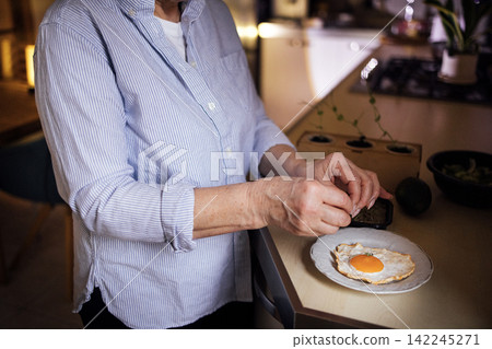 Senior woman in blue striped shirt prepares fried egg on a white plate in a modern kitchen with countertop and cooking utensils visible in the background Senior woman in blue striped shirt prepares fried egg on a white plate in a modern kitchen with countertop and cooking utensils visible in the background 142245271