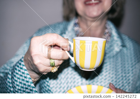 Mature woman holding a yellow striped teacup with a matching bowl, wearing a patterned blouse and jewelry, smiling in a softly lit indoor setting Mature woman holding a yellow striped teacup with a matching bowl, wearing a patterned blouse and jewelry, smiling in a softly lit indoor setting 142245272