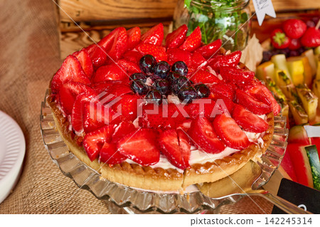 Delicious fresh fruit tart topped with vibrant glazed red strawberries and dark blueberries on a glass cake stand Delicious fresh fruit tart topped with vibrant glazed red strawberries and dark blueberries on a glass cake stand 142245314