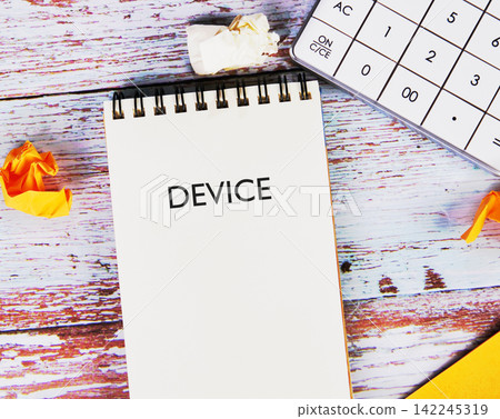 DEVICE word text written on a spiral notepad placed on a wooden desk next to a calculator 142245319