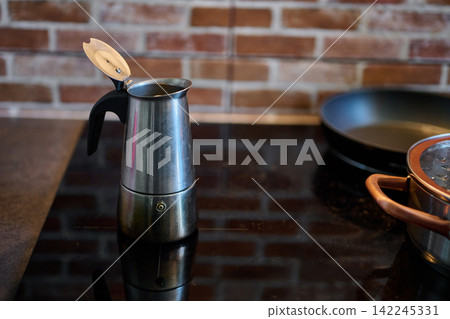 Open silver moka pot standing on a black induction stovetop next to a frying pan and saucepan against a brick wall Open silver moka pot standing on a black induction stovetop next to a frying pan and saucepan against a brick wall 142245331