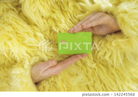 Person wearing a fluffy yellow coat holds a green credit card with a chip, hands positioned in front of a textured background of the coat Person wearing a fluffy yellow coat holds a green credit card with a chip, hands positioned in front of a textured background of the coat 142245568