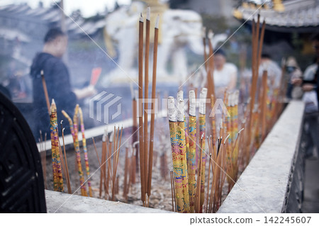 Incense sticks burning in a stone holder at a temple, with blurred figures of worshippers and a statue visible in the background Incense sticks burning in a stone holder at a temple, with blurred figures of worshippers and a statue visible in the background 142245607