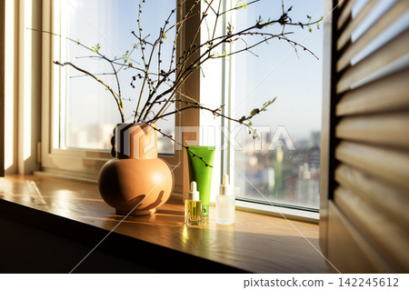 Decorative vase with branches, skincare products, and sunlight streaming through a window on a wooden windowsill in a modern interior setting Decorative vase with branches, skincare products, and sunlight streaming through a window on a wooden windowsill in a modern interior setting 142245612