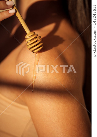 Close-up of honey dripping from wooden dipper onto shoulder of a woman with smooth skin illuminated by soft natural light in a serene indoor setting Close-up of honey dripping from wooden dipper onto shoulder of a woman with smooth skin illuminated by soft natural light in a serene indoor setting 142245613