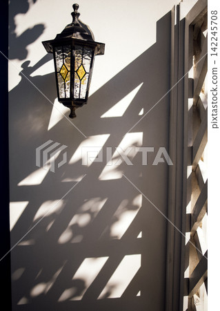 Vintage lantern with stained glass design casting intricate shadows on a white wall beside a decorative wooden lattice structure in a bright outdoor setting Vintage lantern with stained glass design casting intricate shadows on a white wall beside a decorative wooden lattice structure in a bright outdoor setting 142245708
