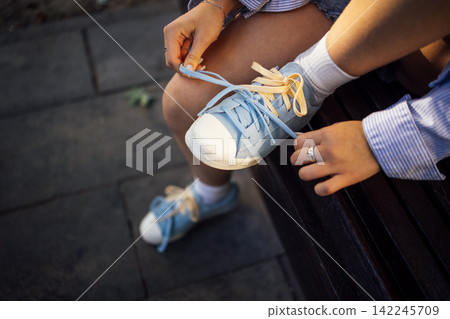 Female wearing blue sneakers ties shoelaces while sitting on a wooden bench, sunlight illuminating her hands and casual outfit in an outdoor setting Female wearing blue sneakers ties shoelaces while sitting on a wooden bench, sunlight illuminating her hands and casual outfit in an outdoor setting 142245709
