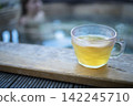 Clear glass cup filled with yellow beverage placed on wooden surface beside hot tub with blurred figure in background, relaxing in warm water 142245710