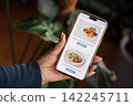 Hand holding smartphone displaying food delivery app with images of two dishes, one with tacos and the other with a salad, surrounded by indoor plants 142245711