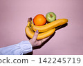Hand holding three fruits including bananas, an orange, and a green apple against a pink background, showcasing vibrant colors and fresh produce 142245719