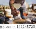 Hand of a male bartender holding a margarita glass with lime garnish, another cocktail glass on a table in a sunny outdoor lounge setting 142245723