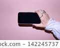 Hand holding a smartphone with a blank screen against a pink background, showcasing a modern device suitable for mockup designs and digital presentations 142245737