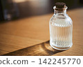 Clear glass bottle with cork stopper containing liquid, placed on wooden surface, casting a shadow in warm natural light 142245770