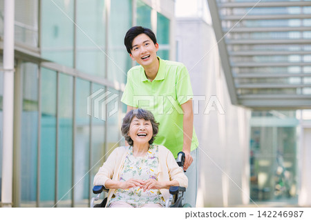 一位年輕的男性護理員推著輪椅,旁邊是一位老年婦女。 一位年輕的男性護理員推著輪椅,旁邊是一位老年婦女。 142246987