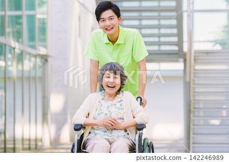 A young male caregiver pushing a wheelchair and an elderly woman A young male caregiver pushing a wheelchair and an elderly woman 142246989