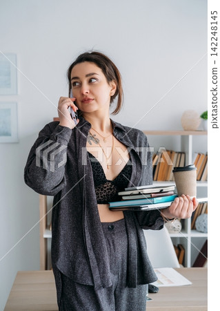 Office manager. Digital workflow. Female talking on smartphone with notebook coffee using laptop preparing for business paperwork. Office manager. Digital workflow. Female talking on smartphone with notebook coffee using laptop preparing for business paperwork. 142248145