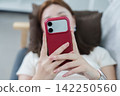 Close up of woman holding red smartphone while relaxing at home 142250560