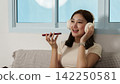 Asian woman wearing headphones sending voice message on smartphone at home 142250581