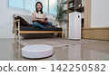 Robot vacuum cleaner operating on living room floor while woman rests on sofa 142250582