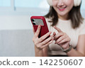 Asian woman using smartphone and wearing headphones at home for digital entertainment 142250607