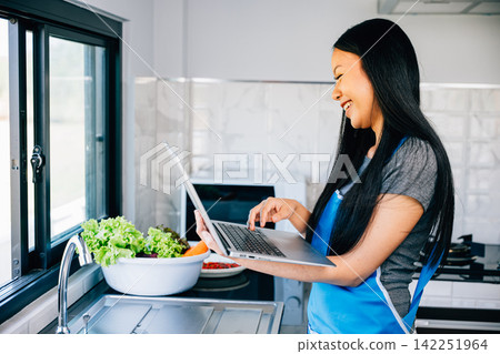 A woman stands in the kitchen happily cooking while using a laptop for video tutorials A woman stands in the kitchen happily cooking while using a laptop for video tutorials 142251964