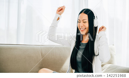 Happy woman in headphones holds phone dances and gestures on sofa Happy woman in headphones holds phone dances and gestures on sofa 142251988