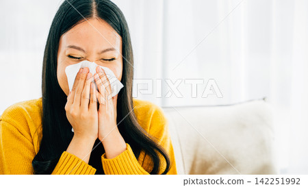 Asian woman blowing her nose holding a handkerchief experiencing flu symptoms at home 142251992